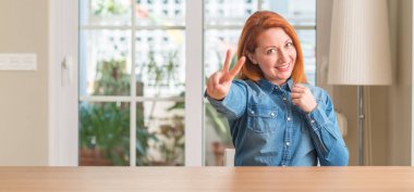 Gülümseyen Kızıl saçlı kadın evde parmak zafer işareti yaparken gösterilen kamera arıyorum. İki numaralı.