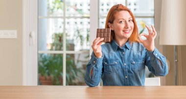 Parmak, mükemmel sembolü ile ok işareti yapıyor evde çikolata bar tutan Kızıl saçlı kadın