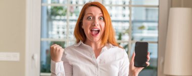 Kızıl saçlı kadın tezahürat duygu heyecan gurur çığlık ve zafer ve başarı çok kutluyor, smartphone evde kullanma