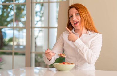 Kızıl saçlı kadın yeme hububat ile ahududu eliyle işaret çok mutlu ev ve yan parmak