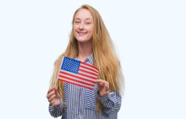 Amerika Birleşik Devletleri bayrağı ile ayakta ve diş gösterilen kendine güvenen bir gülümsemeyle gülümseyerek mutlu bir yüz tutan sarışın genç kadın