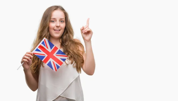 年轻的金发女子持有英国国旗惊讶的想法或问题指着一个快乐的脸 — 图库照片