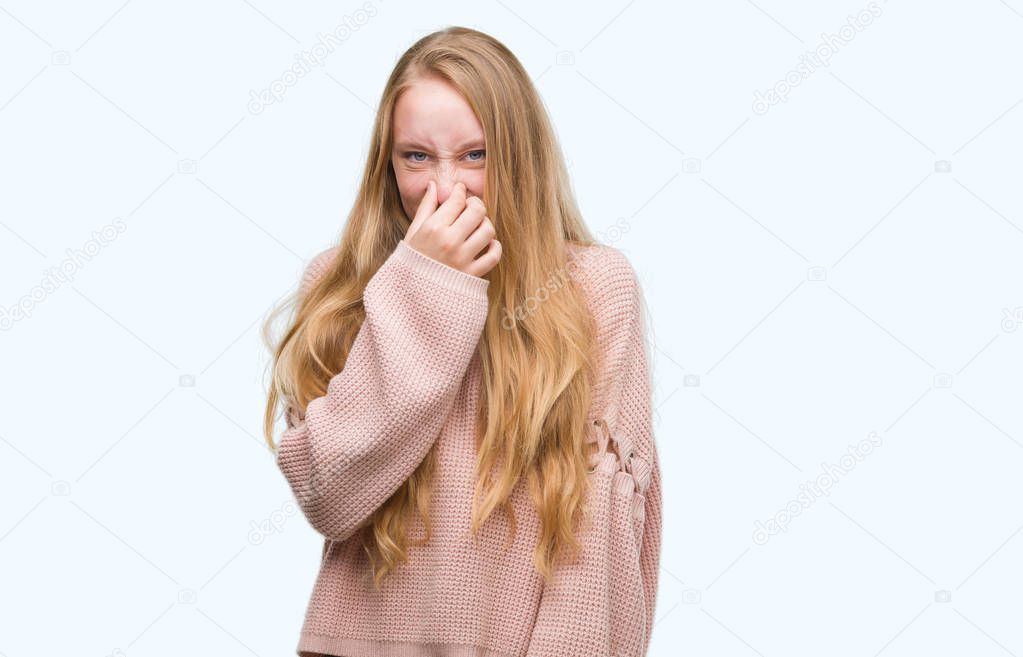Blonde teenager woman wearing pink sweater smelling something stinky and disgusting, intolerable smell, holding breath with fingers on nose. Bad smells concept.