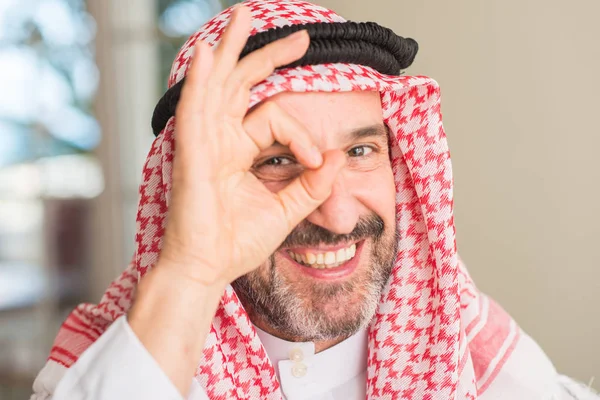 Middle age arabian man at home with happy face smiling doing ok sign with hand on eye looking through fingers