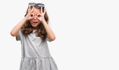 Esmer Latin kız dilini, parmaklarının arasından baktığını yapışmasını dürbün gibi ok jest yapıyor güneş gözlüğü takıyor. Deli ifade.