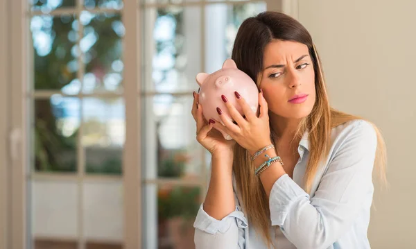 Beautiful young woman wondering how much money has into piggy bank at home. Business concept.
