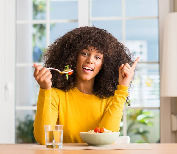 家でパスタのサラダを食べるアフリカ系アメリカ人の女性を幸せな顔で ナンバーワンのアイデアや質問ポインティング指で驚かせた — ストック写真