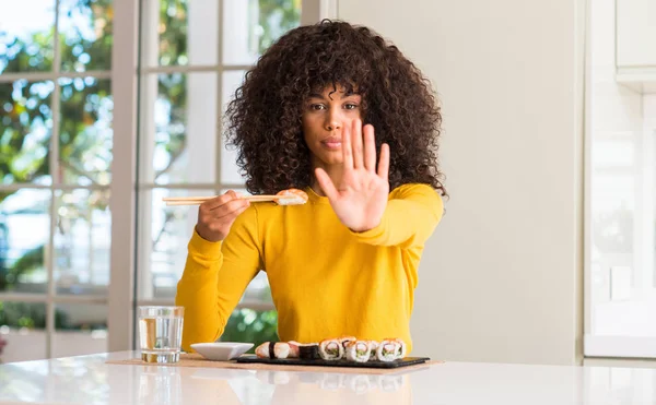 African american woman eating sushi using chopsticks at home with open hand doing stop sign with serious and confident expression, defense gesture