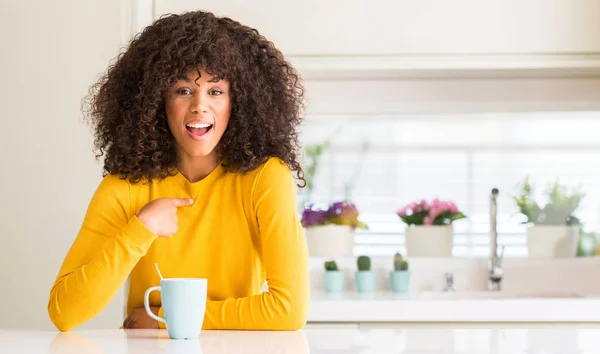美丽的非洲裔美国妇女拿着一杯咖啡在家里与惊讶的脸指向自己的手指 — 图库照片