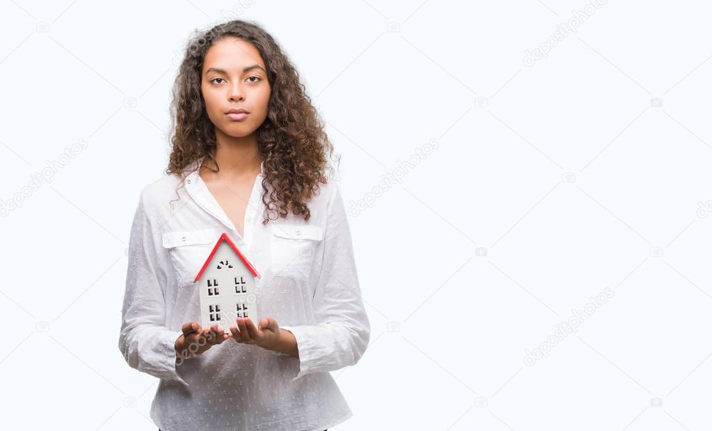 Young hispanic real state agent woman holding small house with a confident expression on smart face thinking serious
