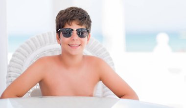 Tatil günlerinde ayakta ve diş gösterilen kendine güvenen bir gülümsemeyle gülümseyerek mutlu bir yüz ile plajda bir sandalyede oturan genç çocuk