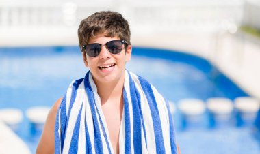 Tatil günlerinde genç çocuk ayakta ve diş gösterilen kendine güvenen bir gülümsemeyle gülümseyerek mutlu bir yüz ile plajda yüzme havuzunda