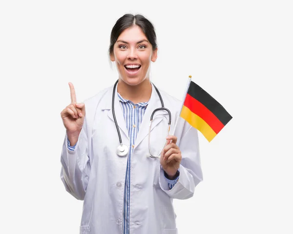 年轻的西班牙裔医生妇女持有德国国旗惊讶的想法或问题指着一个快乐的脸 — 图库照片