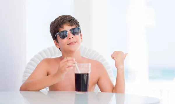 Young child on holidays drinking soda pointing and showing with thumb up to the side with happy face smiling