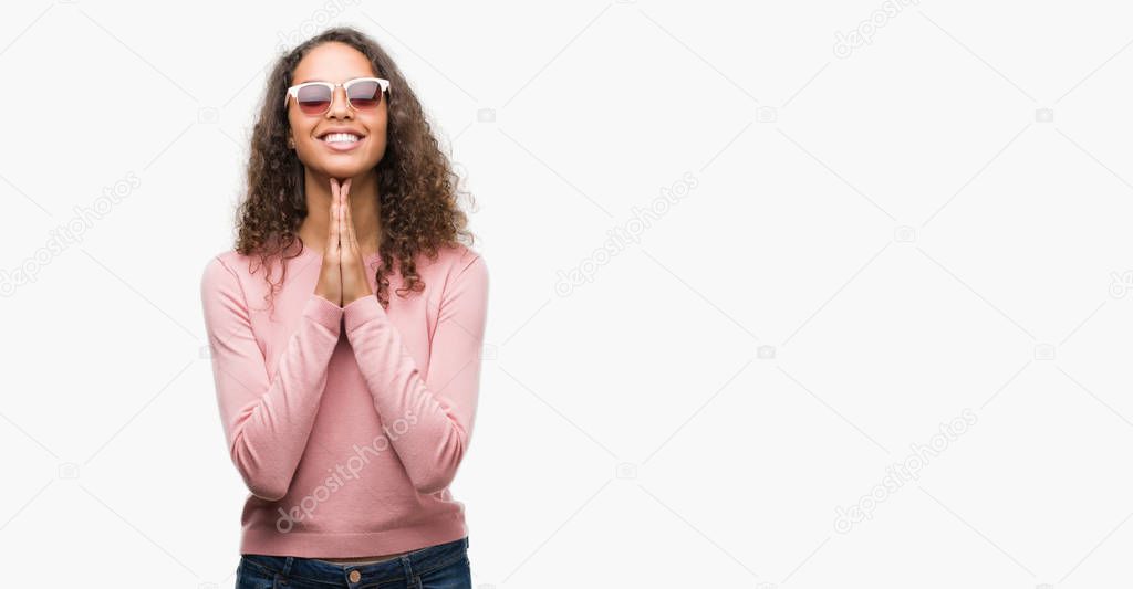 Beautiful young hispanic woman wearing sunglasses praying with hands together asking for forgiveness smiling confident.