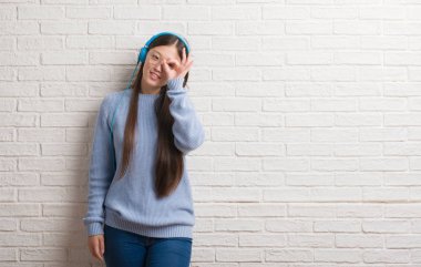 Mutlu yüz el parmaklarının arasından seyir göz üzerinde ok işaretiyle yapıyor gülümseyen ile kulaklıklar giyen tuğla duvarın üzerinden Genç Çinli kadın
