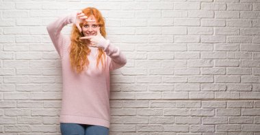 Elle yapma çerçeve ve parmakları ile mutlu yüz gülümseyen tuğla duvarın üzerinden duran genç Kızıl saçlı kadın. Yaratıcılık ve Fotoğrafçılık kavramı.