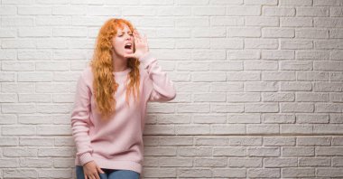 Bağırarak ve ağzı üzerinde el ile yan için çığlık tuğla duvarın üzerinden duran genç Kızıl saçlı kadın. İletişim kavramı.