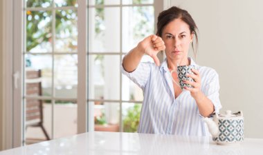 Middle aged woman drinking a cup of tea or coffee with angry face, negative sign showing dislike with thumbs down, rejection concept clipart