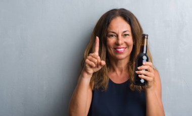 Bira şişesi sürpriz bir fikir ile tutarak gri grunge duvarın üzerinden duran yaş İspanyol kadın orta veya gülen yüz, bir numara ile işaret parmağı soru