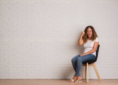 Sandalye üzerinde beyaz tuğla walll rahatsız ve öfke, çılgın ve yükseltilmiş eliyle diye bağıran ile sinirli bağırarak öfke kavramı oturan orta yaş İspanyol kadın