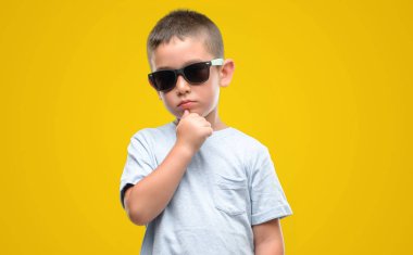 Koyu saçlı küçük çocuk güneş gözlüğü ciddi bir yüz düşünme hakkında soru, çok karışık fikir giyiyor