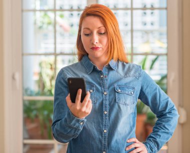 Kızıl saçlı kadın evde Smartphone akıllı yüzünde ciddi düşünme emin bir ifade ile kullanma