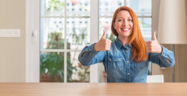 Redhead woman at home success sign doing positive gesture with hand, thumbs up smiling and happy. Looking at the camera with cheerful expression, winner gesture. clipart