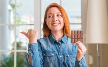 El ve parmak ile yukarı dönük mutlu yüz gülümseyen ile evde çikolata bar tutan Kızıl saçlı kadın