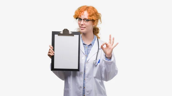 年轻红发医生的女人拿着剪贴板做 标志与手指 优秀的符号 — 图库照片