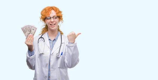 Young Redhead Doctor Woman Holding Dollars Pointing Showing Thumb Side — Stock Photo, Image