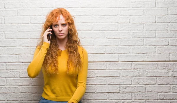 Young redhead woman standing over brick wall talking on the phone with a confident expression on smart face thinking serious