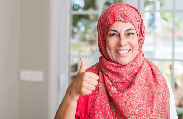 Middle aged muslim woman wearing hijab happy with big smile doing ok sign, thumb up with fingers, excellent sign