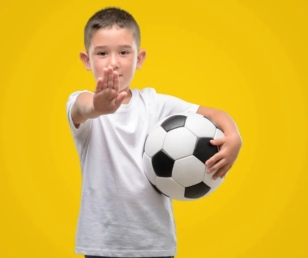 Dark haired little child playing with soccer ball with open hand doing stop sign with serious and confident expression, defense gesture