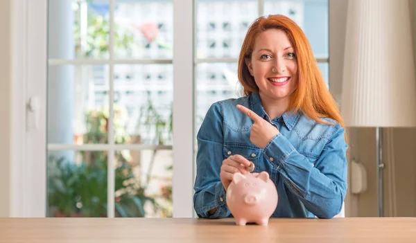 Rödhårig Kvinna Sparar Pengar Piggy Bank Hem Mycket Nöjda Pekar — Stockfoto