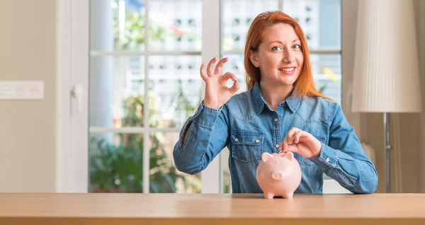 Redhead Woman Saves Money Piggy Bank Home Doing Sign Fingers — Stock Photo, Image