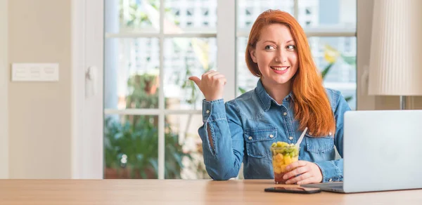 红头发的女人用电脑笔记本在家里吃水果指着手和手指向上微笑的笑脸 — 图库照片
