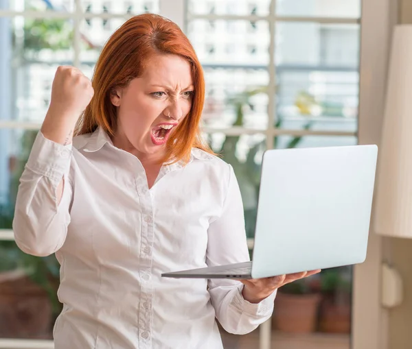 Redhead woman using computer laptop at home annoyed and frustrated shouting with anger, crazy and yelling with raised hand, anger concept