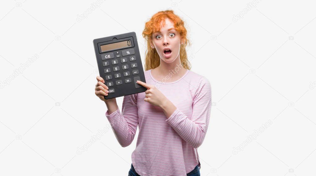 Young redhead woman holding big calculator scared in shock with a surprise face, afraid and excited with fear expression