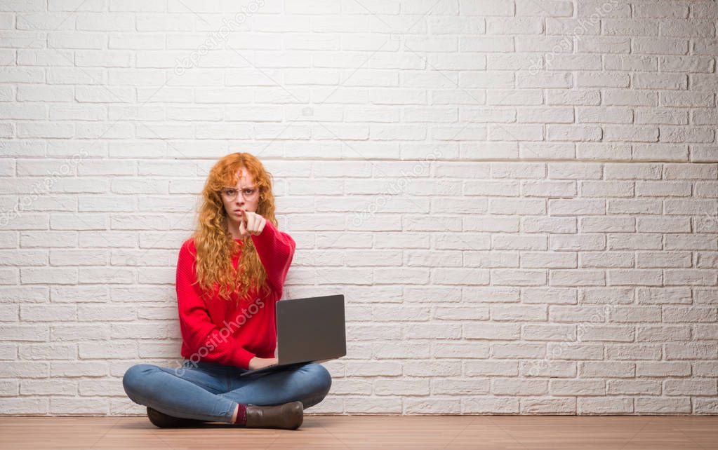 Young redhead woman sitting over brick wall using computer laptop pointing with finger to the camera and to you, hand sign, positive and confident gesture from the front
