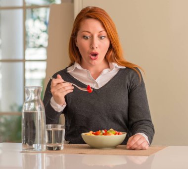 Kızıl saçlı kadın evde meyve tabağı, kivi ve çilek yemek korkmuş bir sürpriz yüz ile şok korkmuş ve heyecanlı korku ifadesi ile