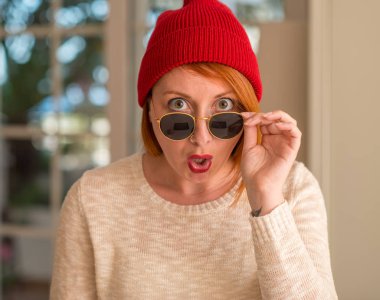 Yün şapka ve güneş gözlüğü bir sürpriz yüz, korkuyor ve heyecanlı korku ifadesi ile şok korkmuş giymiş şık Kızıl saçlı kadın