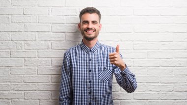 Genç yetişkin adam beyaz tuğla duvardan mutlu büyük gülümseme ok işareti, başparmak ile parmak, mükemmel işareti yapıyor ayakta