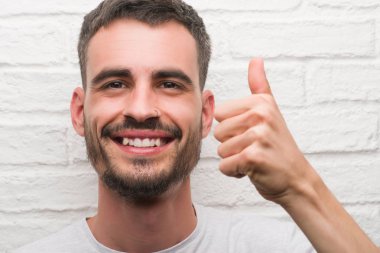Genç yetişkin adam beyaz tuğla duvardan mutlu büyük gülümseme ok işareti, başparmak ile parmak, mükemmel işareti yapıyor ayakta