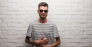 Genç yetişkin adam beyaz tuğla duvarın mide üzerinde el ile üzerinden çünkü ayakta güneş gözlüğü takıyor hazımsızlık, ağrılı hastalık hasta hissediyor. Ağrısı kavramı.