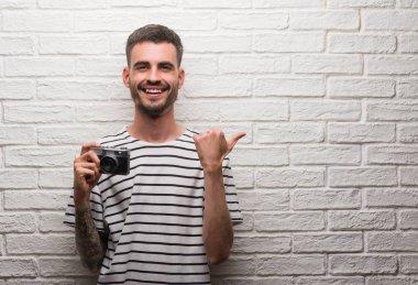 Genç adam işaret ve başparmak kadar mutlu yüz gülümseyen tarafı ile gösterilen beyaz tuğla duvarın üzerinden vintage fotoğraf makinesi ayakta tutan