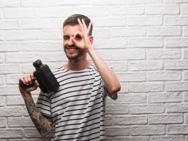 Genç adam Holding video kamera ile el parmaklarının arasından seyir göz üzerinde ok işaretiyle yapıyor gülümseyen mutlu yüz beyaz tuğla duvarı ayakta filme