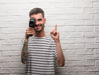 Genç adam filme tuğla duvar bir fikirle sürpriz beyaz video kamera ayakta tutan veya gülen yüz, bir numara ile işaret parmağı soru