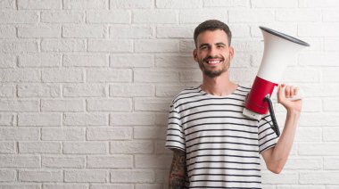 Genç yetişkin adam ayakta ve diş gösterilen kendine güvenen bir gülümsemeyle gülümseyerek mutlu bir yüz ile megafon holding