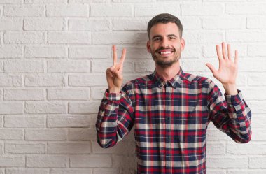 Gösterilen ve parmakları ile işaret beyaz tuğla duvarın üzerinden duran genç yetişkin adam sırasında kendinden emin ve mutlu gülümseyerek yedi numara.